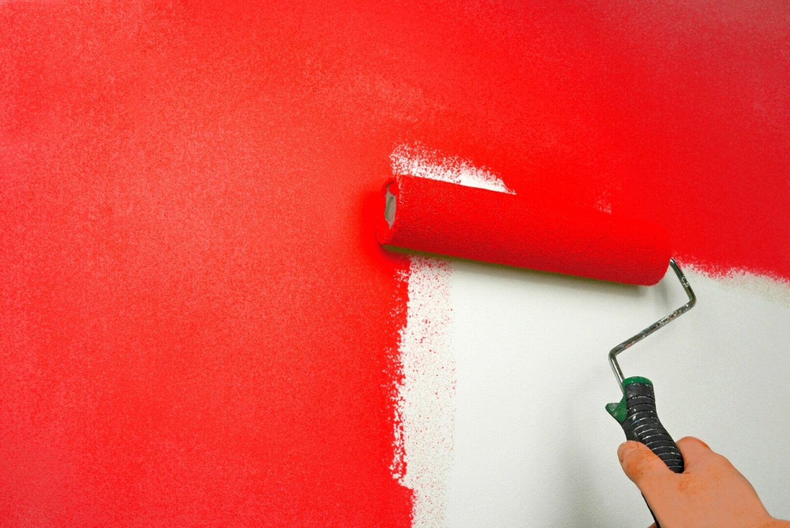 A hand using a red paint roller to apply a fresh coat on a partially painted white wall.